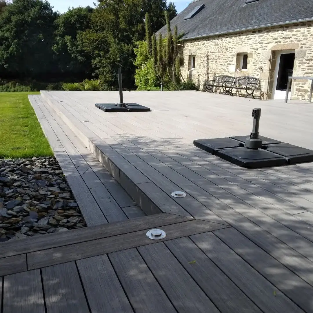 Grande terrasse en bois avec spots LED intégrés réalisée par Agencement Lennon devant une maison en pierre.